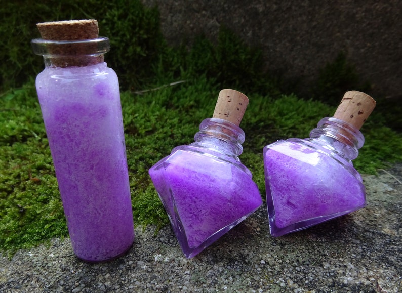 May include: Three glass bottles with cork stoppers filled with a purple and white glittery liquid. The bottles are on a mossy green surface.