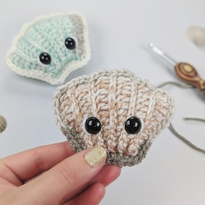 Puede incluir: Concha de ganchillo hecha a mano con ojos de botón negros. La concha es de color beige y gris, sostenida por una mano con una uña con purpurina dorada. Otra concha está en el fondo, en color azul claro. El proyecto de artesanía está sobre una superficie blanca.