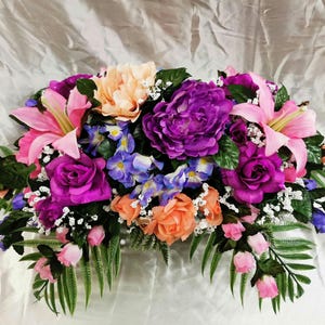 May include: A floral arrangement featuring pink lilies, purple peonies, peach roses, and purple and blue flowers. The arrangement is set on a white background.