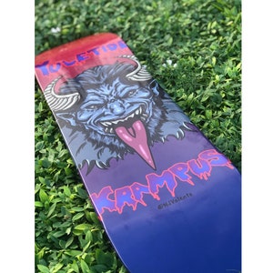 May include: A purple and blue skateboard deck with a graphic of a blue demon with a pink tongue and the text "YULETIDE KARMUS" in pink and blue lettering. The text "@NJValente" is in white lettering at the bottom of the deck.