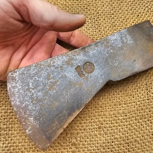 May include: A vintage axe head with a worn, silver finish. The axe head has a rounded edge and a small circular stamp with the number "2000" on the side.