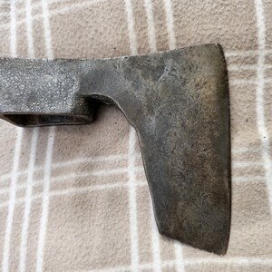 Könnte beinhalten: Ein verwitterter, antiker Axtkopf mit rechteckigem Auge und geschwungener Klinge. Das Metall hat ein dunkles, gealtertes Aussehen, was auf eine lange Nutzung hindeutet. Der Axtkopf liegt auf hellem, gemustertem Stoff.