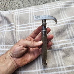 Könnte beinhalten: Ein Vintage-Klauenhammer mit gebogener Klaue und Metallkopf. Der Hammer hat einen dunklen, strukturierten Griff und wird in einer Hand gehalten. Der Hammer liegt auf einer beige-weiß karierten Decke.