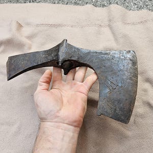 May include: An antique axe head with a curved blade and a pointed top. The metal has a dark, aged appearance with visible wear. The axe head is held in a hand, showing its size and shape. The axe head is likely made of iron or steel.