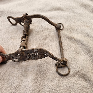 Pode incluir: Um antigo freio de cavalo de metal enferrujado com um design articulado. O freio apresenta uma série de elos conectados e um bocal curvo com uma borda dentada. O freio foi projetado para ser colocado na boca de um cavalo para controlá-lo.