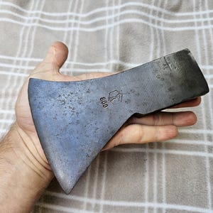 Op de afbeelding: Een donkergrijze metalen bijlkop met een verweerd, getextureerd oppervlak. Het metaal is voorzien van een gestempeld zeshoekig logo met een gestileerd symbool en het getal 800. De bijlkop heeft een brede, gebogen snede en een platte achterkant.
