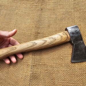 Peut inclure: Une hache à manche en bois avec une tête en métal noir. La hache a une légère courbure de la lame et repose sur une surface en toile de jute brune.