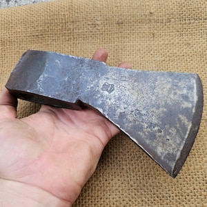 Può includere: Una testa di ascia vintage con una finitura in metallo grigio usurato. La testa dell'ascia ha una leggera curva sulla lama e un bordo arrotondato. La testa dell'ascia ha un piccolo numero stampato sul lato.
