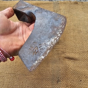 Op de afbeelding: Een roestige metalen bijlkop met een gebogen steel. De bijlkop heeft een klein, driehoekig logo met de tekst "125H" en "3" erboven.