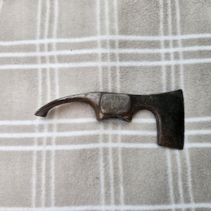 Puede incluir: Una herramienta de metal oscuro y desgastado que presenta una punta curva en un lado y una hoja rectangular plana en el otro. La sección central tiene una abertura rectangular y la superficie muestra signos de oxidación y antigüedad.