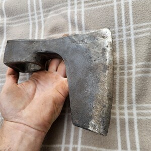 以下が含まれることがあります： 幅広の刃と柄の穴がある、風化したグレーの金属製の斧頭。 斧頭には使用感があり、刃先はわずかに丸みを帯びています。 格子柄の背景に、手に持たれた道具です。
