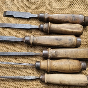 Puede incluir: Seis formones antiguos con mangos de madera y hojas de acero de diferentes formas y tamaños. Los formones están dispuestos sobre una superficie texturizada de color beige. Herramientas probablemente utilizadas para trabajar la madera o tallar.