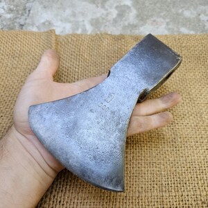 Könnte beinhalten: Ein verwitterter Axtkopf mit gebogener Klinge und dunkler Metalloberfläche. Der Axtkopf wird in einer Hand gehalten, um seine Größe und Form zu zeigen. Der Axtkopf hat die Nummer "000" eingestanzt.