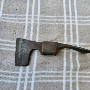 Op de afbeelding: Een vintage, roestige metalen bijl met een gebogen handvat en een rechthoekig blad. De bijlkop is donkergrijs van kleur en vertoont tekenen van ouderdom en gebruik. De handgreep is gebogen.