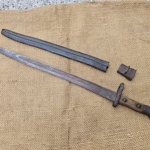 Puede incluir: Una bayoneta antigua con mango de madera y hoja de metal, acompañada de una funda de cuero negro y una pequeña trabilla de cuero. La bayoneta se muestra sobre una superficie de arpillera beige.