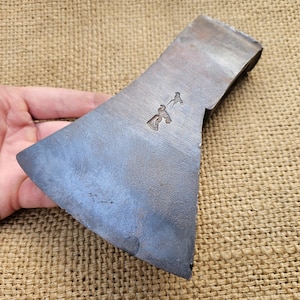 Könnte beinhalten: Ein blauer Stahl-Axtkopf mit einem kleinen, geprägten Design auf der Oberfläche. Der Axtkopf liegt auf einer Jutestoff-Oberfläche.