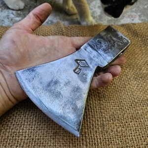 Puede incluir: Una cabeza de hacha de metal vintage con un acabado plateado desgastado. La cabeza del hacha se sostiene en una mano, mostrando su afilada hoja y las marcas en relieve. La cabeza del hacha descansa sobre una superficie tejida marrón.