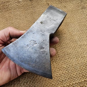 May include: A vintage steel axe head with a worn, textured surface. The axe head has a curved blade and a rectangular eye for the handle. The metal has a dark, aged appearance, with markings near the eye.