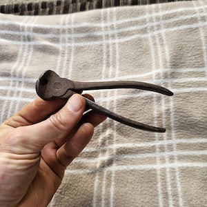Può includere: Un utensile in metallo arrugginito e vintage con manici curvi e una testa arrotondata. L'utensile è tenuto in mano su uno sfondo a quadri beige e bianco. L'utensile sembra essere un tipo di pinza.