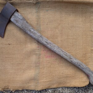 以下が含まれることがあります： 木製の柄と金属製のヘッドを持つヴィンテージの斧。斧は茶色の麻布の上に置かれています。