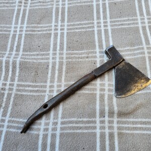 Op de afbeelding: Een kleine, antieke bijl met een donkere, verweerde uitstraling. De metalen kop heeft een driehoekig blad en een hamer aan de andere kant. De steel is gebogen met een gat aan het uiteinde. De bijl ligt op een geruit oppervlak.