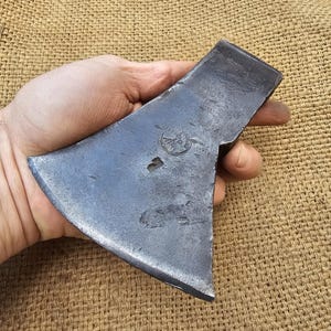 Pode incluir: Uma cabeça de machado vintage forjada à mão com uma lâmina de aço e um acabamento cinza desgastado. A cabeça do machado tem uma pequena marca em forma de coração na lâmina.
