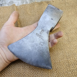 Pode incluir: Uma cabeça de machado de aço vintage com uma lâmina larga, segurada em uma mão. A cabeça do machado tem uma forma ligeiramente curva e é marcada com o número "1000". A cabeça do machado é de cor cinza prateado e mostra sinais de desgaste.