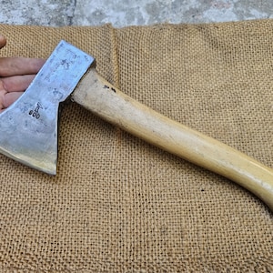 Può includere: Un'ascia vintage con manico in legno e lama in acciaio. La testa dell'ascia è contrassegnata con il numero "600". Il manico è marrone chiaro e la lama è argentata. L'ascia è appoggiata su una superficie di juta.