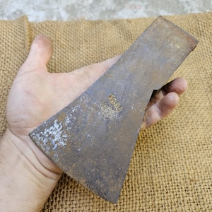 Pode incluir: Uma cabeça de machado vintage com uma aparência enferrujada e envelhecida. A lâmina de metal tem uma forma triangular com uma borda ligeiramente curva. Um pequeno emblema circular é visível perto do topo. A cabeça do machado é segurada em uma mão, em uma superfície de juta.