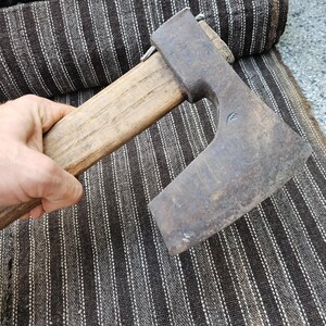 May include: A hand axe featuring a weathered, dark gray metal head with a wide, curved blade edge and a straight, light brown wooden handle. The metal head shows signs of oxidation and wear, securely attached to the wooden shaft with a metal wedge.