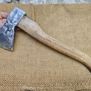 Può includere: Un'ascia vintage con manico in legno e testa in metallo. La testa dell'ascia ha una lama leggermente curva ed è contrassegnata dal numero "600". Il manico è di colore marrone chiaro e l'ascia è appoggiata su una superficie di juta.