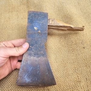Könnte beinhalten: Eine Vintage-Axt mit einem Holzstiel. Der Axtkopf ist aus Metall und hat eine abgenutzte, rostige Oberfläche. Der Axtkopf ist mit dem Text "Dan 1950" geprägt.