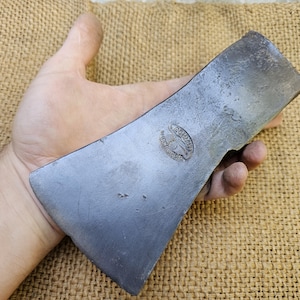 Könnte beinhalten: Ein verwitterter Axtkopf mit metallischem Finish. Der Axtkopf wird in einer Hand gehalten, was seine Größe und Form zeigt. Der Axtkopf hat ein rundes Emblem mit Text. Der Hintergrund ist eine strukturierte, beige Oberfläche.
