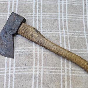 Puede incluir: Un hacha de mano con una cabeza de metal desgastada y un mango de madera. La cabeza de metal presenta un logotipo estampado y el número 1000. La cabeza del hacha muestra signos de oxidación y desgaste, mientras que el mango tiene una textura natural.