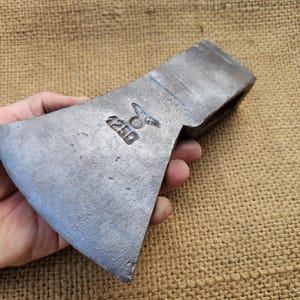 Peut inclure: Une tête de hache vintage avec un manche en métal. La tête de hache est en acier et a une lame légèrement incurvée. La tête de hache est marquée du numéro "220" et d'un symbole qui ressemble à un oiseau stylisé.