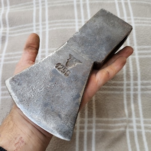 Pode incluir: Uma cabeça de machado de metal vintage com uma superfície desgastada e texturizada. A cabeça do machado tem uma lâmina larga e um olho retangular para o cabo. O número "1250" e um emblema estão estampados. A cabeça do machado é segurada em uma mão.