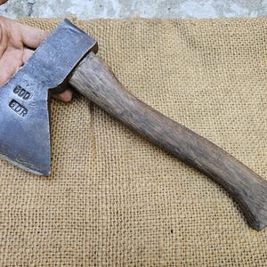 May include: A vintage axe with a dark metal head and a wooden handle. The axe head has the stamped text "800 GDR". The handle is a dark brown colour. The axe is resting on a tan burlap surface.