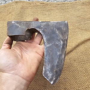 Pode incluir: Uma mão segura uma cabeça de machado grande, robusta, de aço com uma lâmina curva. A cabeça do machado é cinza e tem uma superfície áspera e texturizada.