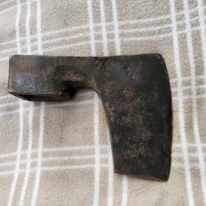 Op de afbeelding: Een vintage bijlkop met een donkere, verweerde uitstraling. Het metalen gereedschap heeft een rechthoekig oog voor de steel en een gebogen blad. De bijlkop ligt op een beige en witte geruite stof.