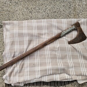 Könnte beinhalten: Eine Vintage-Axt mit Holzgriff und rostigem Metallkopf. Die Axt liegt auf einer beige-weiß karierten Decke. Der Griff ist dunkelbraun und der Kopf aus dunklem, verwittertem Metall.