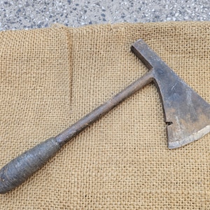 Könnte beinhalten: Eine alte Axt mit einem Metallkopf und einem Holzgriff. Die Axt liegt auf einer Jutefläche und unterstreicht ihren rustikalen Charakter. Die Klinge weist Gebrauchsspuren auf.