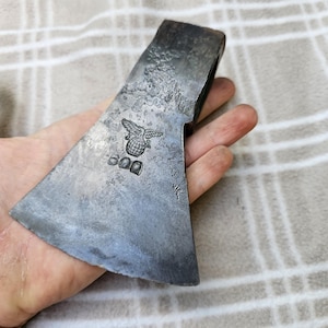 May include: A dark grey metal axe head with a weathered, textured surface. The metal features a stamped emblem of an eagle perched atop a globe, positioned above the number 600. The axe head has a wide, flared blade edge and a thick poll.