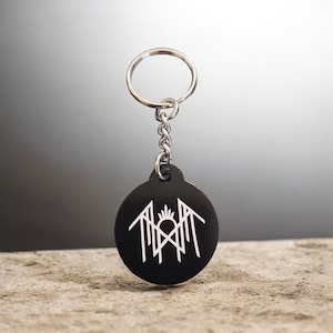 May include: A circular black keychain featuring a white geometric line art design centered on the front. The keychain is attached to a silver-colored metal split ring by a short chain. The design consists of symmetrical angular lines and a central peak.