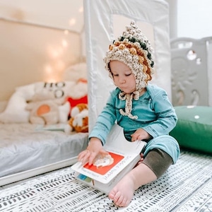 May include: A child wearing a brown, green, and yellow crocheted hat with a pointed top sits on a black and white patterned rug and reads a book titled "Animals Everywhere".