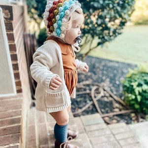 Puede incluir: Un ni&ntilde;o peque&ntilde;o con un c&aacute;rdigan blanco, un vestido marr&oacute;n, calcetines azules hasta la rodilla y zapatos marrones. El ni&ntilde;o lleva un gorro de ganchillo de colores con la parte superior puntiaguda.