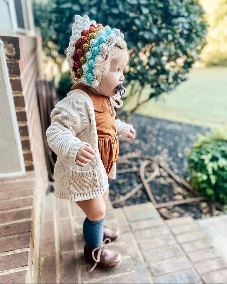 Puede incluir: Un ni&ntilde;o peque&ntilde;o con un c&aacute;rdigan blanco, un vestido marr&oacute;n, calcetines azules hasta la rodilla y zapatos marrones. El ni&ntilde;o lleva un gorro de ganchillo de colores con la parte superior puntiaguda.