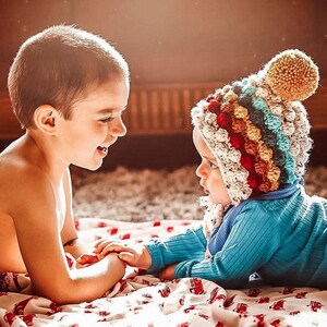 Puede incluir: Dos ni&ntilde;os, uno con un gorro de crochet de colores, est&aacute;n tumbados en una manta roja y blanca. Se miran y sonr&iacute;en.