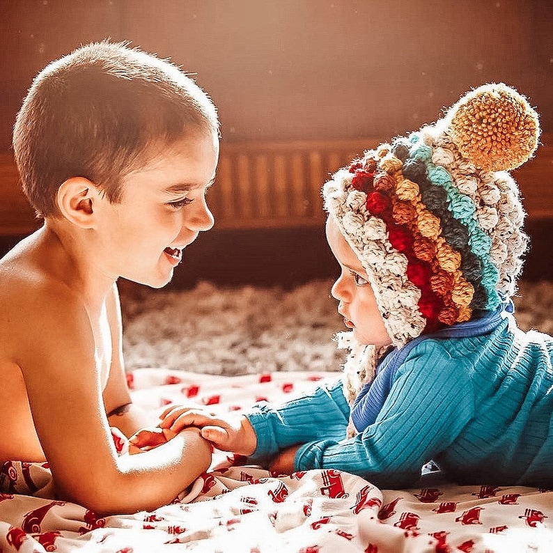 Puede incluir: Dos ni&ntilde;os, uno con un gorro de crochet de colores, est&aacute;n tumbados en una manta roja y blanca. Se miran y sonr&iacute;en.