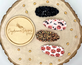 May include: Four Halloween-themed hair clips on a wooden background. The clips feature a black glitter clip, a pink and black clip with bats and pumpkins, an orange and black glitter clip, and a white clip with orange pumpkins.