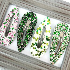 May include: Four hair clips with a scalloped edge. Each clip is decorated with green glitter and shamrock designs. The clips are arranged in a row on a white surface.